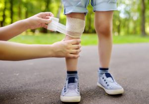A parent bandages the knee of an injured child standing in a park