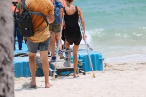 A group of people on the beach. One of them is using crutches.