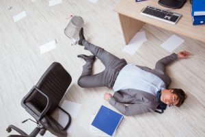 A man in a suit lying unconscious on an office floor surrounded by paper and a toppled over chair.