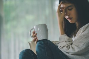A woman sitting against a wall with a coffee mug holding her head